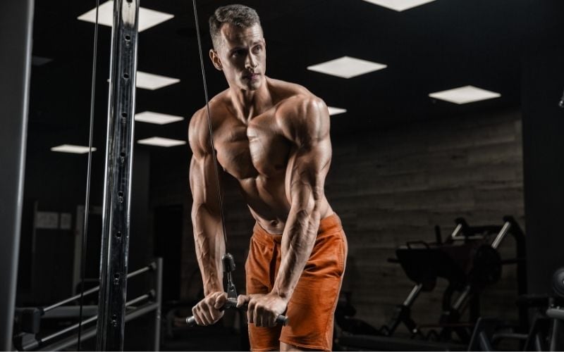 Shirtless man wearing orange shorts doing tricep pushdowns.