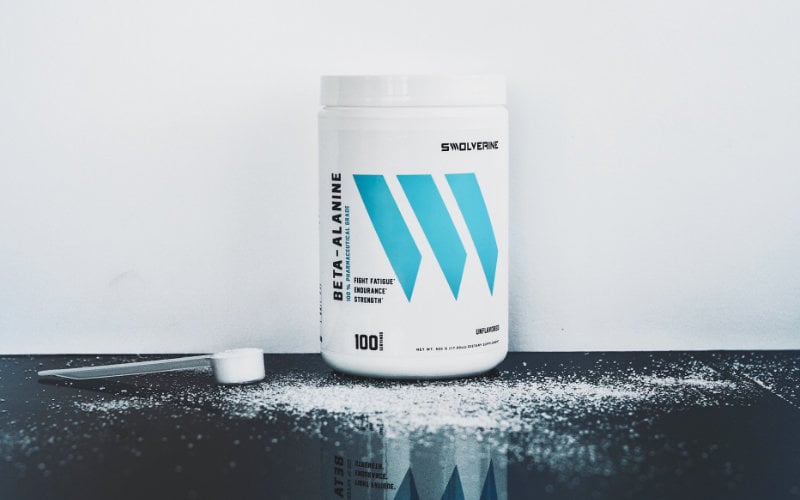 Styled Swolverine Beta-Alanine product shot with beta-alanine bottle in front of white wall and sitting on black reflective surface.