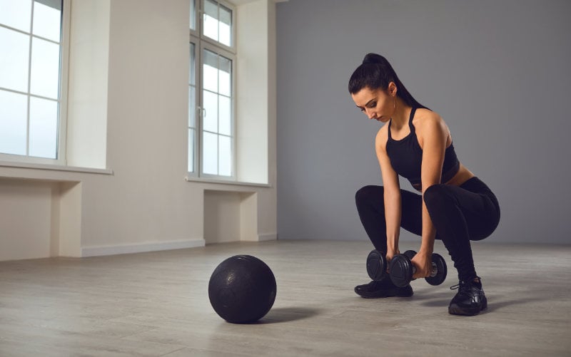 Woman doing dumbbell squats in studio gym with wall ball on the ground in front of her. 