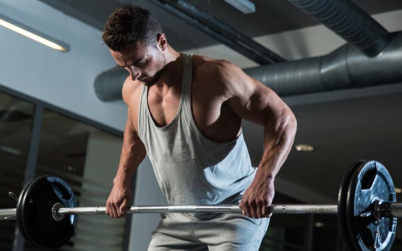 Muscular man wearing a grey stringer doing a barbell row in the gym.