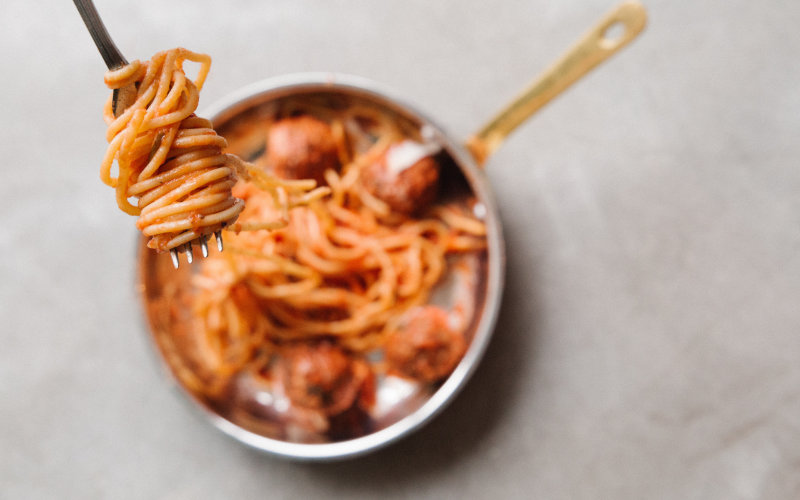 Close up shot of spaghetti on fork with pan of spaghetti and meatballs in the background.