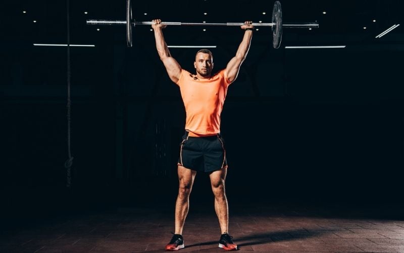 Man in orange shirt holding barbell overhead. 