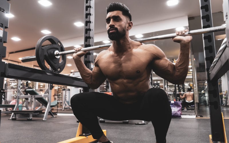 Shirtless man doing barbell back squats in gym. 