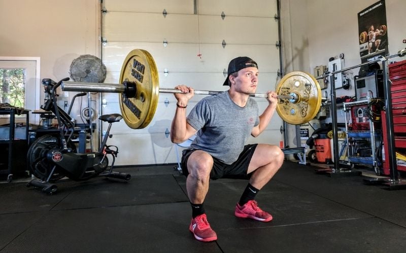 Man in grey t-shirt and black shorts doing a barbell back squat.