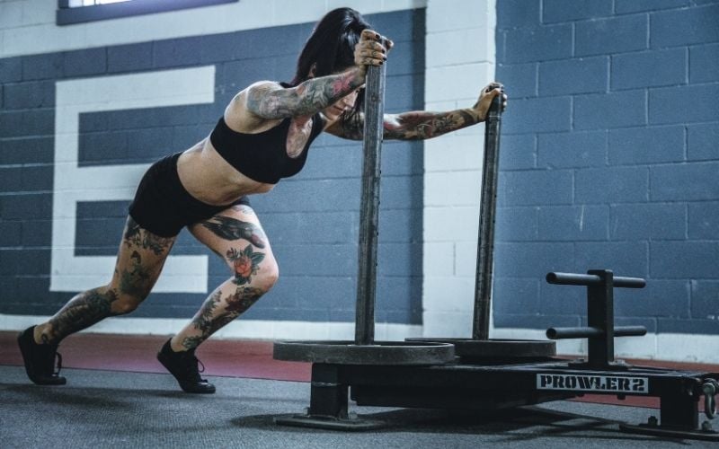 Tattooed muscular woman in black shorts and sports bra pushing a sled.