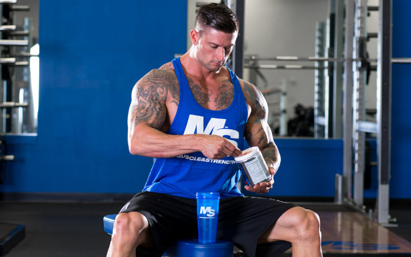 Muscle and Strength model wearing a blue M&S tank and black shorts scooping supplement powder into shaker cup.
