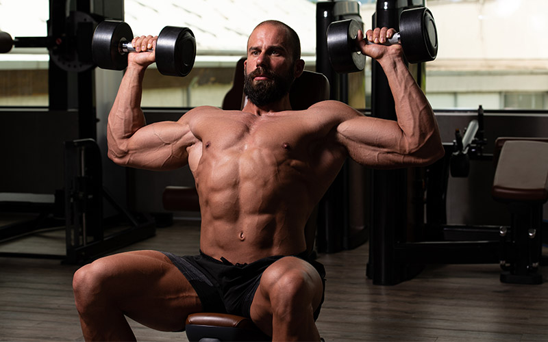 Muscular man in black shorts performing seated dumbbell shoulder press