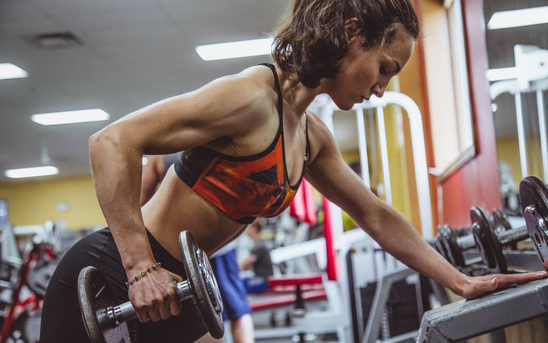 Athletic woman in red sports bra performing a single arm dumbbell row off of a dumbbell rack.
