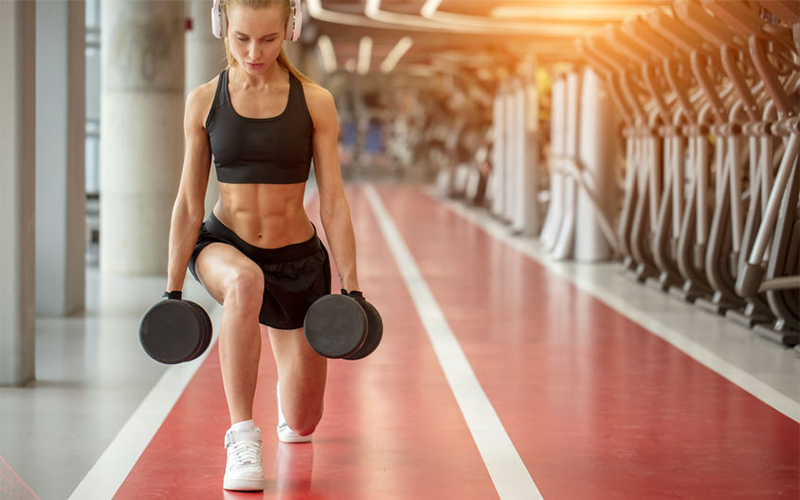 Woman in black sports bra and shorts wearing white headphones doing walking lunges with dumbbells in each hand.